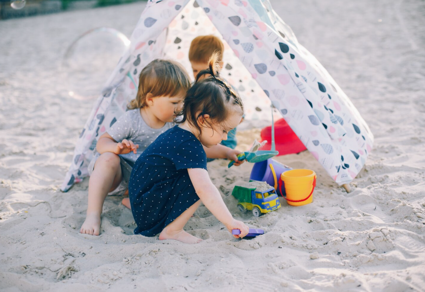 Young children playing in sand near a small, patterned play tent with buckets and spades
