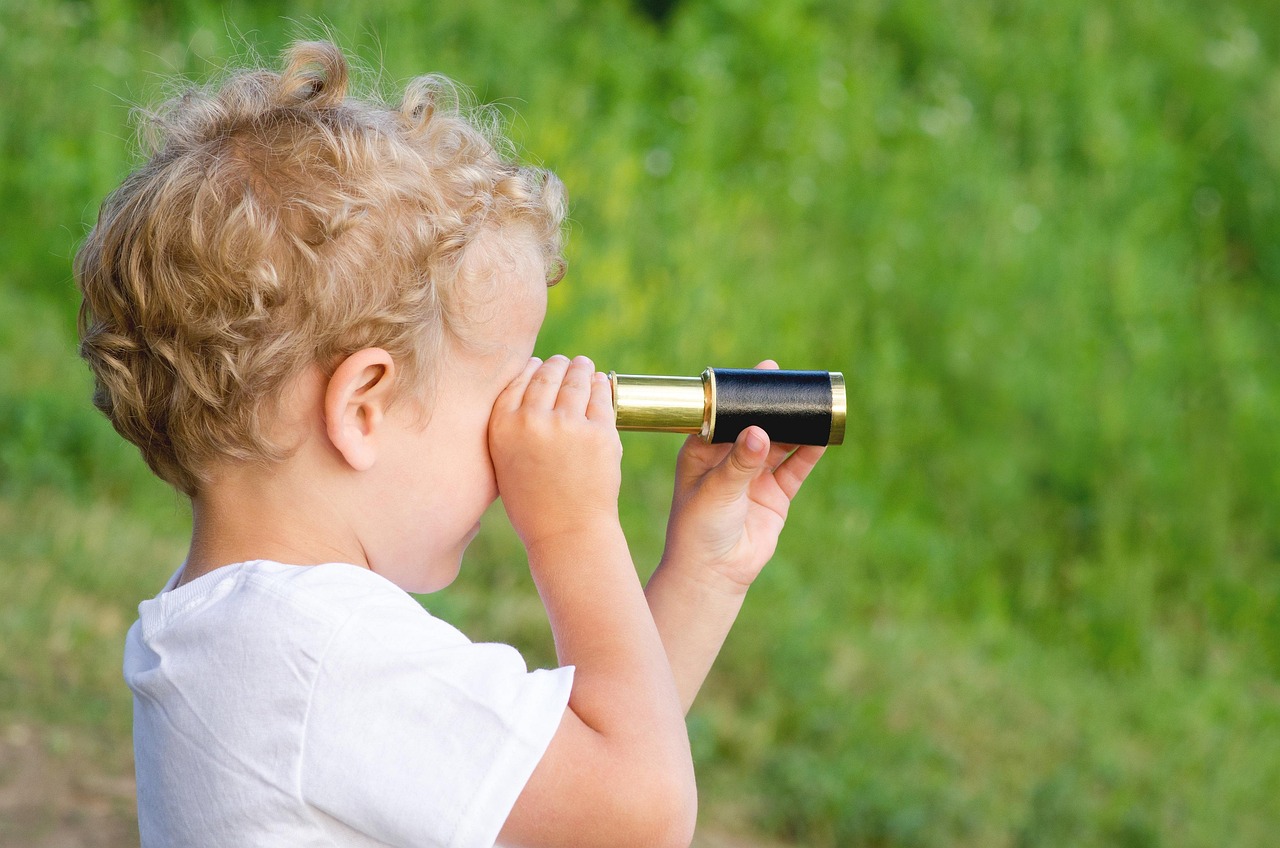 A young child with curly blond hair using a small black-and-gold telescope. Child faces right against a blurred green outdoor background, illustrating exploration or nature observation.