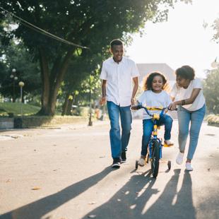 A couple of adults assisting a young child riding a bicycle.