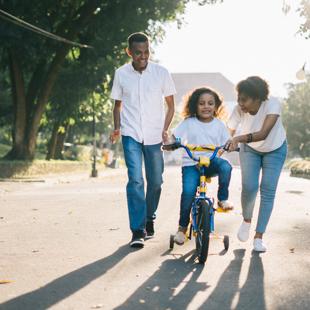 A couple of adults assisting a young child riding a bicycle.
