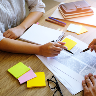 two people collaborating at a wooden table with documents, notebooks, colourful sticky notes, and pens