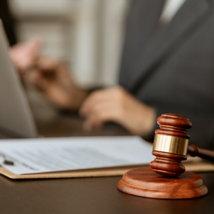 a wooden judge's gavel resting on a desk with a blurred person working on a laptop reviewing documents in the background